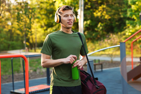 Young man with sports bag, headphones and bottle of water at outdoor gym