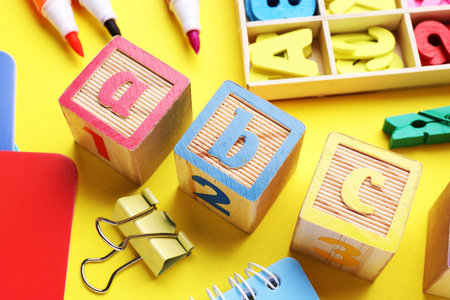 Wooden cubes with letters and school supplies on yellow background, closeup