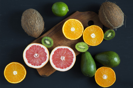 Flat lay. Top view. Sliced colorful  fresh fruits: kiwi, orange, grapefruit and mandarin on wooden board. Coconuts, lime and avocado. Black background. Summer background.の写真素材