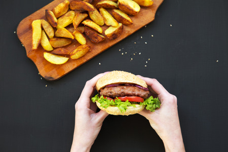 Young girl holds fresh burger. Fried potatoes on wooden board. Top view.の写真素材