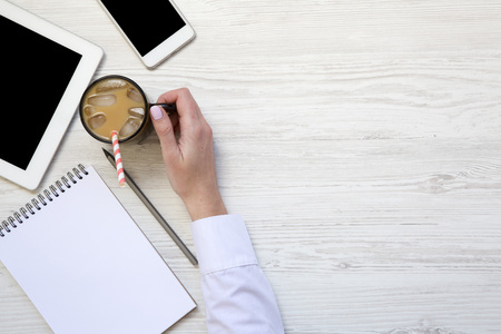 Woman hand holds a cup of ice latte. Feminine workspace with smartphone, latte ice, tablet and notebook. Copy space. Top view. From above. Closeup.の写真素材