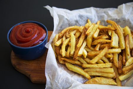 French fries with ketchup on wooden board on dark background, side view, close up