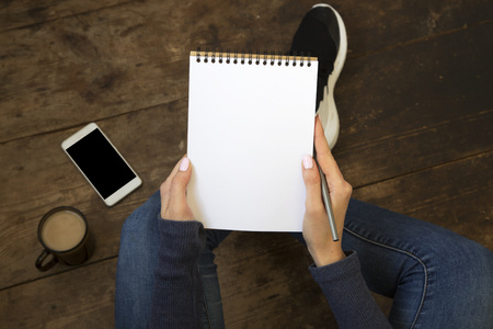 Young girl sits on a wooden floor and holds empty notebook in her hands. Smartphone and coffee on...