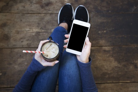 The girl sits on a wooden floor and holds a smartphone with a blank screen and ice latte in her hands. Top view.の写真素材
