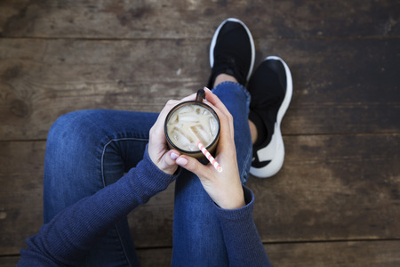 The girl sits on a wooden floor and holds ice latte in her hands. Top view.