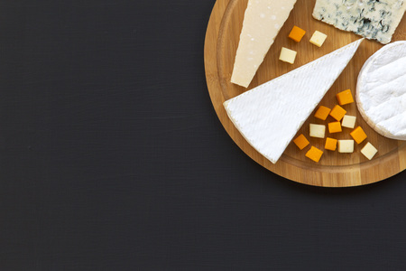 Various types of cheese on round wooden board, top view. Dark background. Space for text. Flat lay....