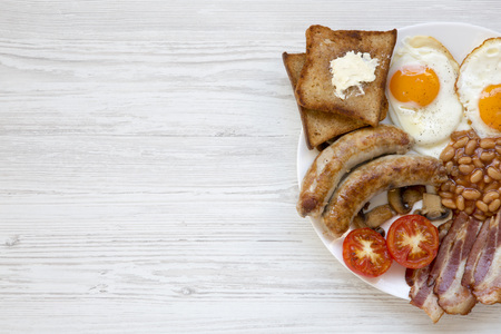 Top view English breakfast with fried eggs, bacon, sausages, beans and toasts. White wooden...