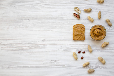 Toast, bowl of peanut butter and peanuts in shells on a white wooden background, top view. Copy...