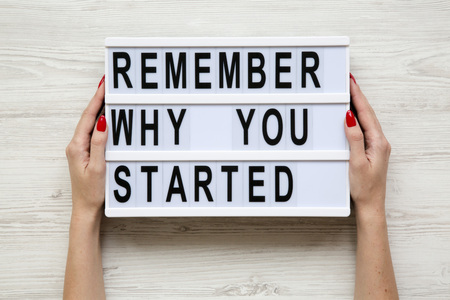Female hands hold modern board with text 'Remember why you started' over white wooden background. From above, flat-lay, overhead.の写真素材