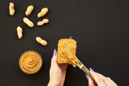 Female hand spreads peanut butter on bread over black background, view from above. Copy space.の写真素材
