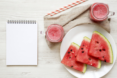 Glass jars of watermelon smoothie, top view. Slices on round plate, drinking straws and blank notepad on white wooden background. Flat lay, from above, overhead.の写真素材