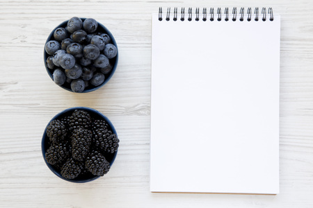 Ripe blackberries and blueberries in bowls with blank notebook over white wooden background, from above. Summer berry. From above, overhead. Copy space.の写真素材