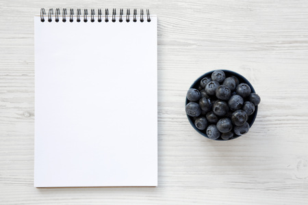 Full bowl of ripe blueberries with notepad over white wooden background, from above. Summer berry. From above, overhead. Copy space.の写真素材