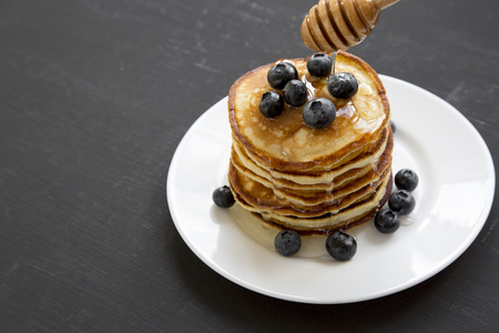 Pancakes with blueberries and honey on a white round plate over black wooden background, side view....