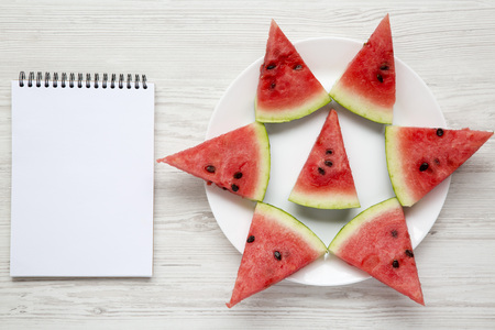 Sliced watermelon on white plate with blank notepad over white wooden background, top view. Flat...