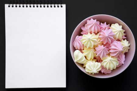 Mini meringues in a pink bowl and blank notepad over black background, top view. Flat lay, overhead, from above.の写真素材