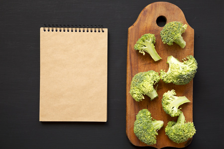 Raw broccoli pieces on rustic wooden board, blank notepad over black background, top view. From...