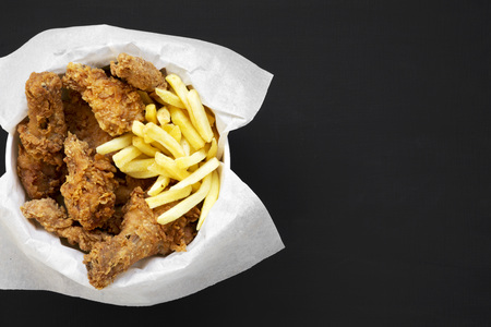 Tasty fastfood: fried chicken drumsticks, spicy wings, French fries and chicken fingers in paper box over black background, top view. Flat lay, overhead, from above. Space for text.の写真素材