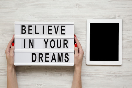 Female hands hold modern board with text 'Believe in your dreams', tablet wit blank screen over white wooden background. From above, overhead, flat-lay.の写真素材