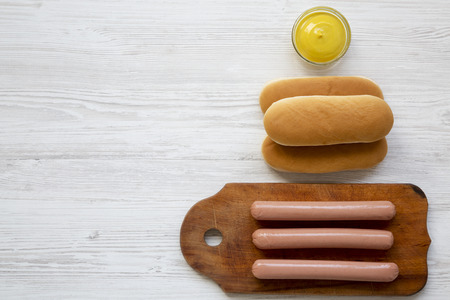 Ingredients for making hot dogs: sausages on wooden board, hot-dog buns and mustard on white wooden surface, overhead view. Flat lay, top view, from above. Copy space.の写真素材