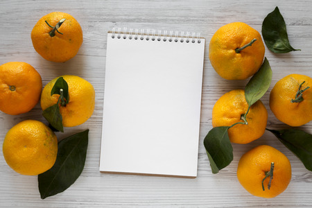 Fresh ripe unshelled tangerines and blank notebook over white wooden background. Flat lay, from above, overhead. Space for text.の写真素材