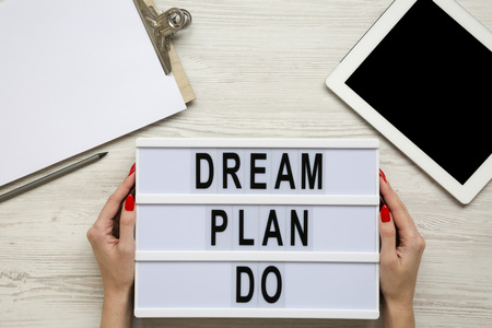 Female hands hold lightbox with text 'Dream plan do', clipboard, pencil and tablet over white wooden...