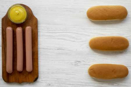 Ingredients for making hotdogs: sausages on wooden board, hot-dog buns and mustard on white wooden background, view from above. Flat lay, overhead, top view.の写真素材
