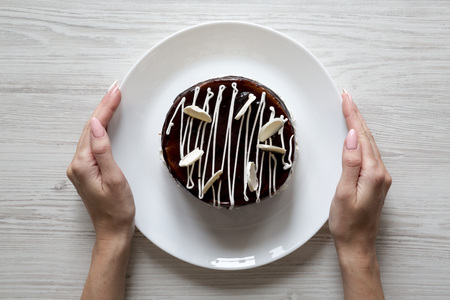 Female hands hold a plate with homemade chocolate cherry cake over white wooden background, top view. Flat lay, overhead, from above.の写真素材