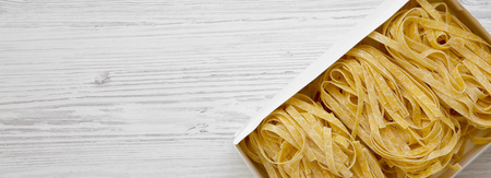 Uncooked pasta fettuccine in paper box on a white wooden surface, top view. Space for text.の写真素材