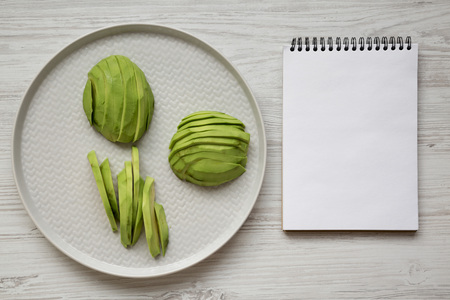 Sliced avocados on gray plate, blank notepad on white wooden table, top view. Overhead, from above,...