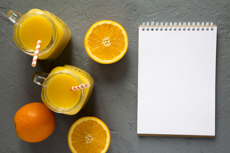 Fresh orange juice in glass jars, blank notepad over gray background, top view. Flat lay, from...