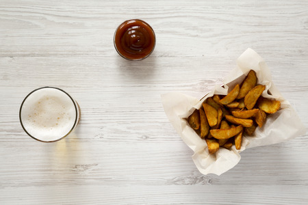 Fried potato wedges in paper box, barbecue sauce and glass of beer on a white wooden table, top...