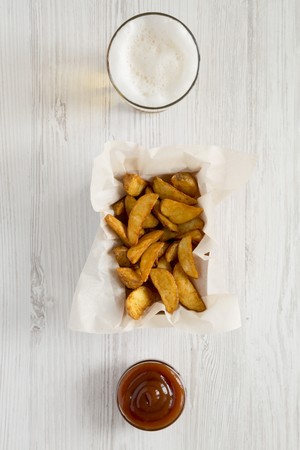 Fried potato wedges in paper box, barbecue sauce and glass of beer on a white wooden surface, top...