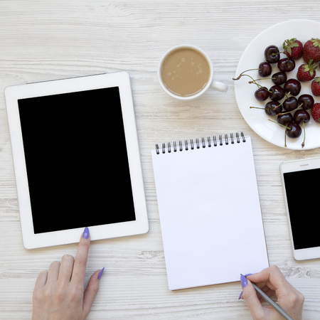 Modern feminine workspace with female hands, smartphone, tablet, notepad, berries and latte over...