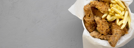 Tasty fastfood: fried chicken legs, spicy wings, French fries and chicken strips in paper box over gray surface, top view. Space for text.の写真素材