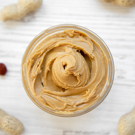 Peanut butter in a bowl and unshelled peanuts on a white wooden surface, top view. From above,...