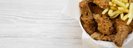 Fast food: fried chicken drumsticks, spicy wings, French fries and chicken strips in paper box over...