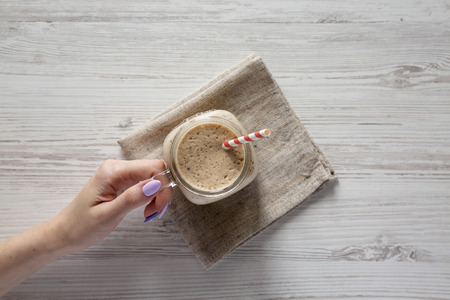 Woman's hand holding a glass jar filled with banana, kiwi, apple smoothie over white wooden surface,...