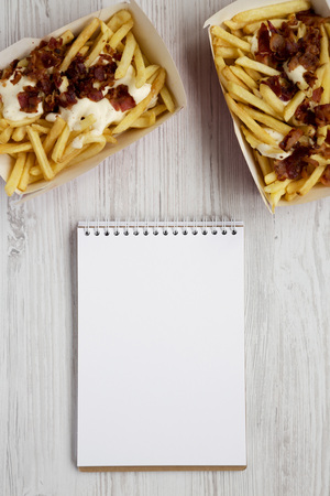 Fast food: french fries with cheese sauce and bacon in paper boxes, blank notepad on a white wooden background, top view.の写真素材
