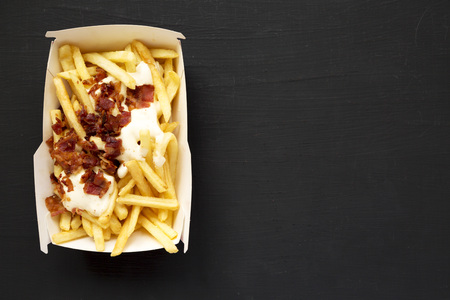 French fries with cheese sauce and bacon in a paper box over black background, top view. Flat lay,...