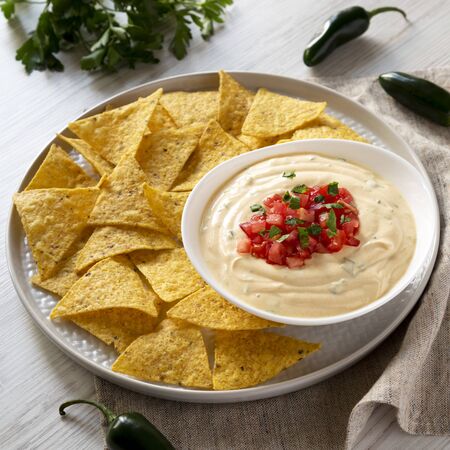 Homemade cheesy dip in a bowl, yellow tortilla chips over white wooden surface, low angle view. Closeup.の写真素材