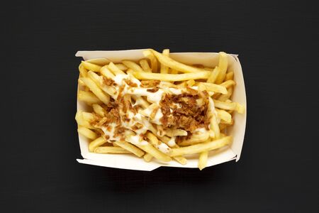 French fries with cheese sauce and onion in a paper box on a black background, top view. Flat lay,...