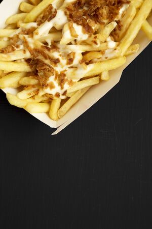 French fries with cheese sauce and fried onion in a paper box on a black background, top view. Flat...