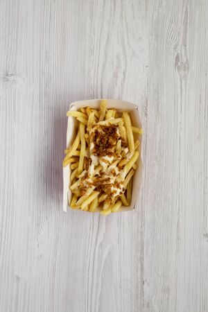 Top view, french fries with cheese sauce and fried onion in a paper box on a white wooden...