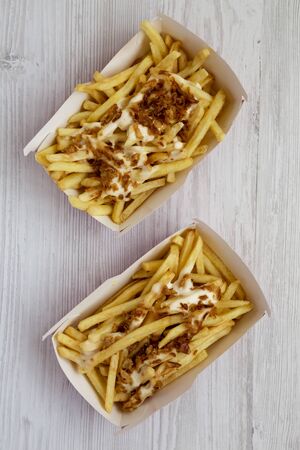 French fries with cheese sauce and fried onion in paper boxes on a white wooden surface, top view....