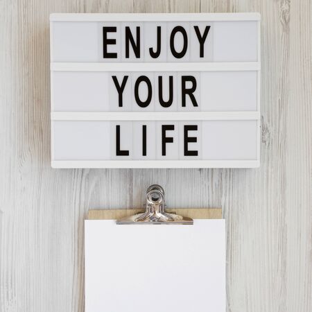 Modern board with text 'Enjoy your life', notepad with blank sheet of paper on a white wooden background, top view. Overhead, from above. Flat lay. Copy space.の写真素材