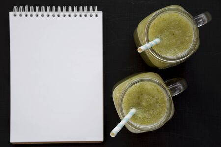 Homemade green cucumber apple smoothie in glass jars, blank notepad on a black background, top view. Flat lay, overhead, from above. Space for text.の写真素材