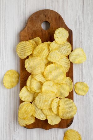 Salted wavy potato chips on a rustic wooden board on a white wooden surface, overhead view. Top...