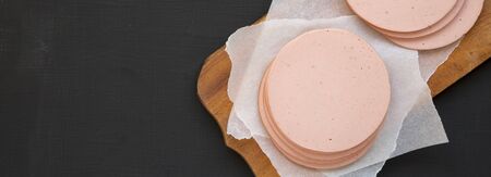 Sliced pork bologna meat on a rustic wooden board on a black background, top view. Flat lay,...