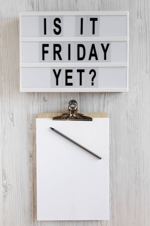 'Is it friday yet?' words on a modern board, clipboard with blank sheet of paper on a white wooden...
